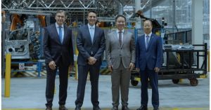 Four men in suits stand in a factory setting with machinery in the background.