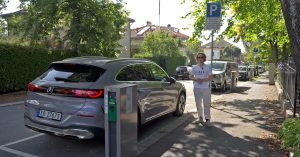 The image shows a silver electric vehicle parked beside a charging station on a street.