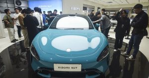 Front view of a blue Xiaomi SU7 in a showroom, surrounded by people.