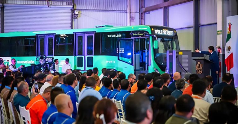 Green electric bus displayed at an event, with audience and podium in a factory setting.