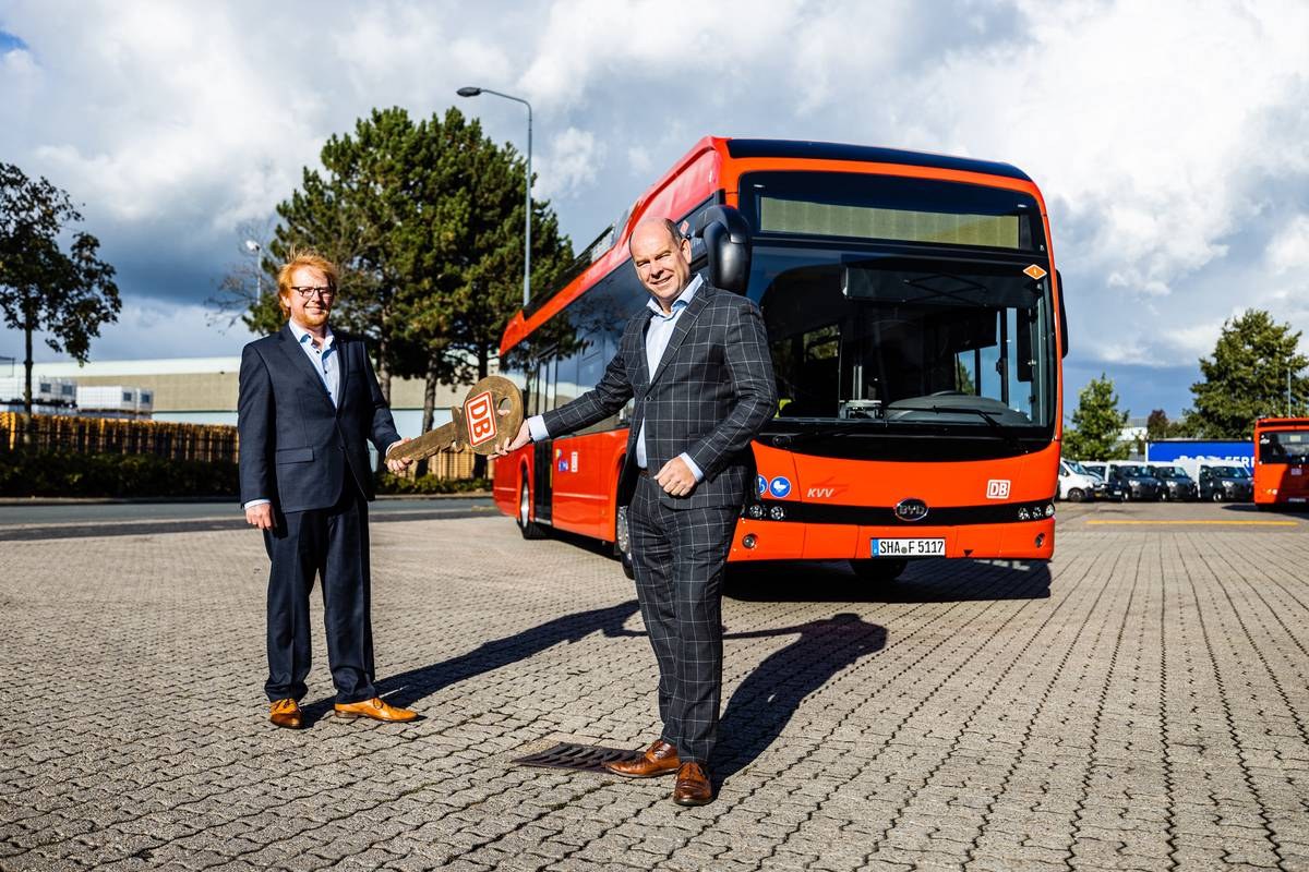 Two men in suits stand in front of a red BYD electric bus, handing over a large key.