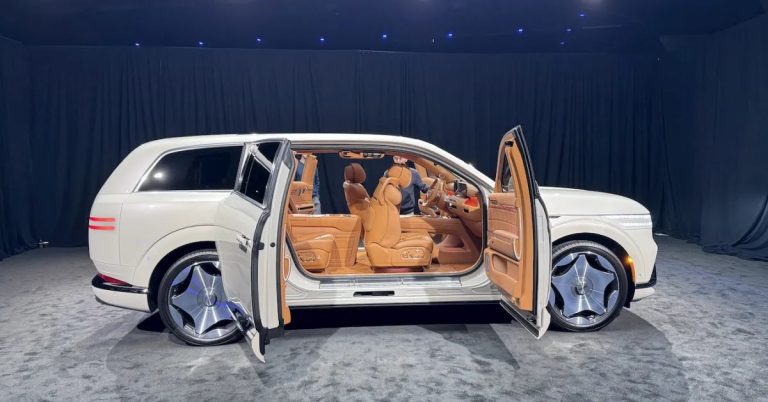 White Genesis GV90 displayed in a showroom, featuring open doors, tan leather seats, and large wheels.