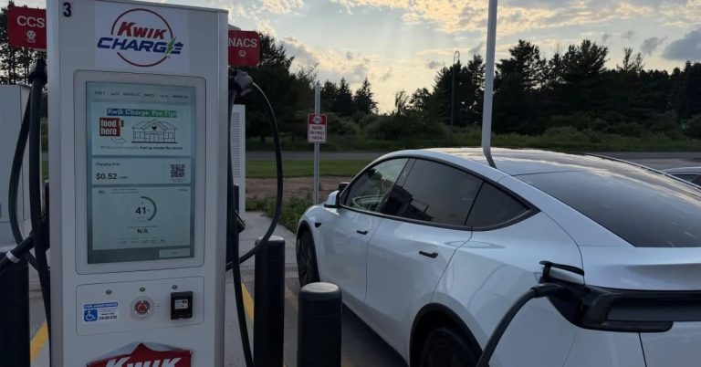 A white electric vehicle is parked at a charging station, with a charging cable connected.