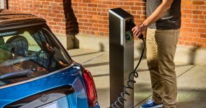 A blue electric vehicle is charging at a station, with a person holding the charging cable.