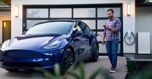 Blue Tesla parked in a driveway, charging port visible, man using smartphone nearby.