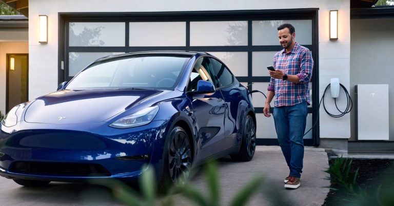 Blue Tesla parked in a driveway, charging port visible, man using smartphone nearby.