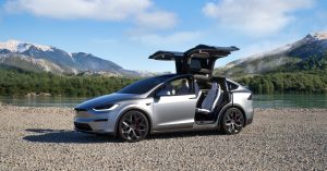 Silver Tesla Model X parked by a lake, featuring falcon-wing doors and sporty wheels.