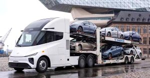 The image shows a white electric truck with a car carrier, parked in an urban setting.