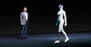 The image features a humanoid robot walking on stage beside a man in a suit.