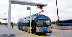 Electric bus parked at charging station, featuring overhead charging arm and digital display.