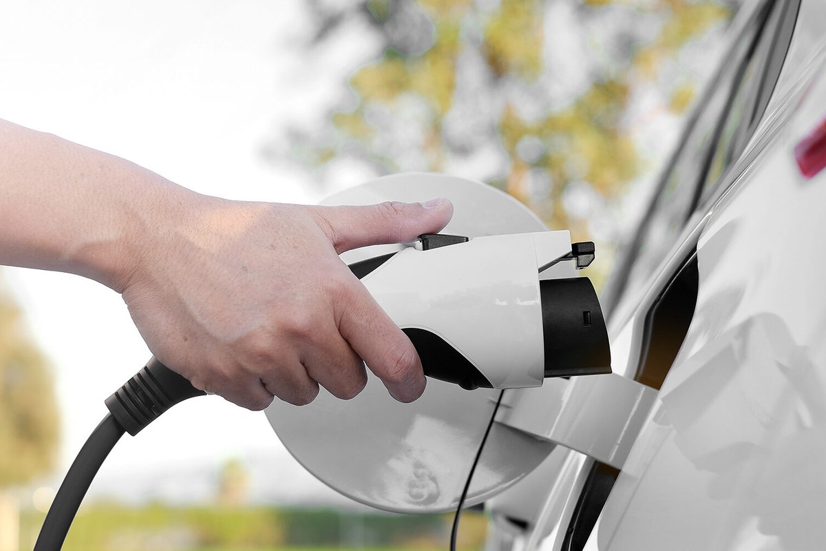 A hand connects a charging plug to an electric vehicle's charging port outdoors.