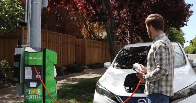 A man stands beside a white EV, connecting a charging cable to its front port.