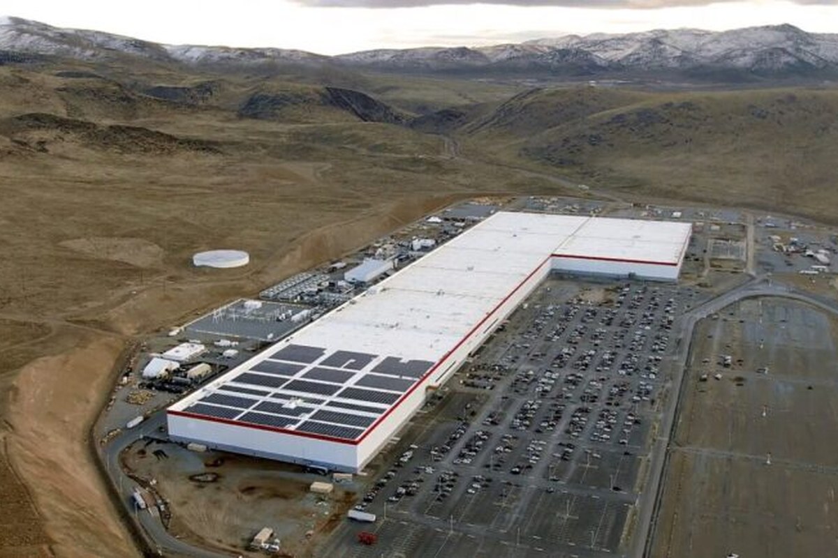 Aerial view of a large factory with solar panels, surrounded by a parking lot and mountains.